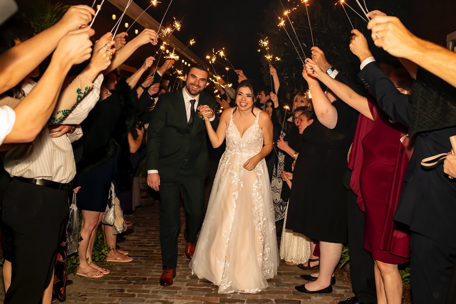 Wedding guests hold sparklers in two rows creating a magical light tunnel exit for the newlyweds at nighttime celebration.
