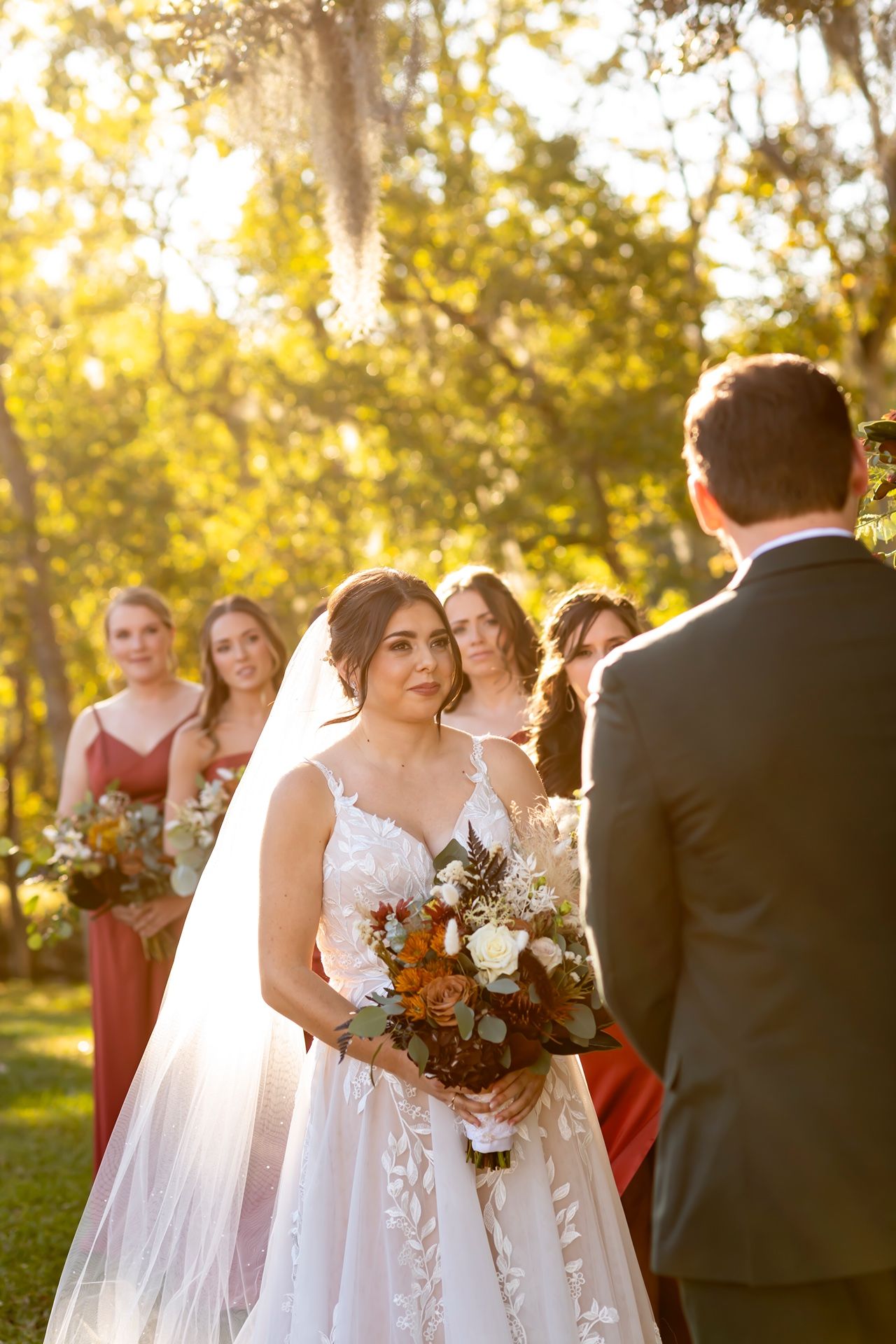 A wedding ceremony in a sunlit outdoor garden setting with bridesmaids in coral dresses and a bride holding a bouquet.