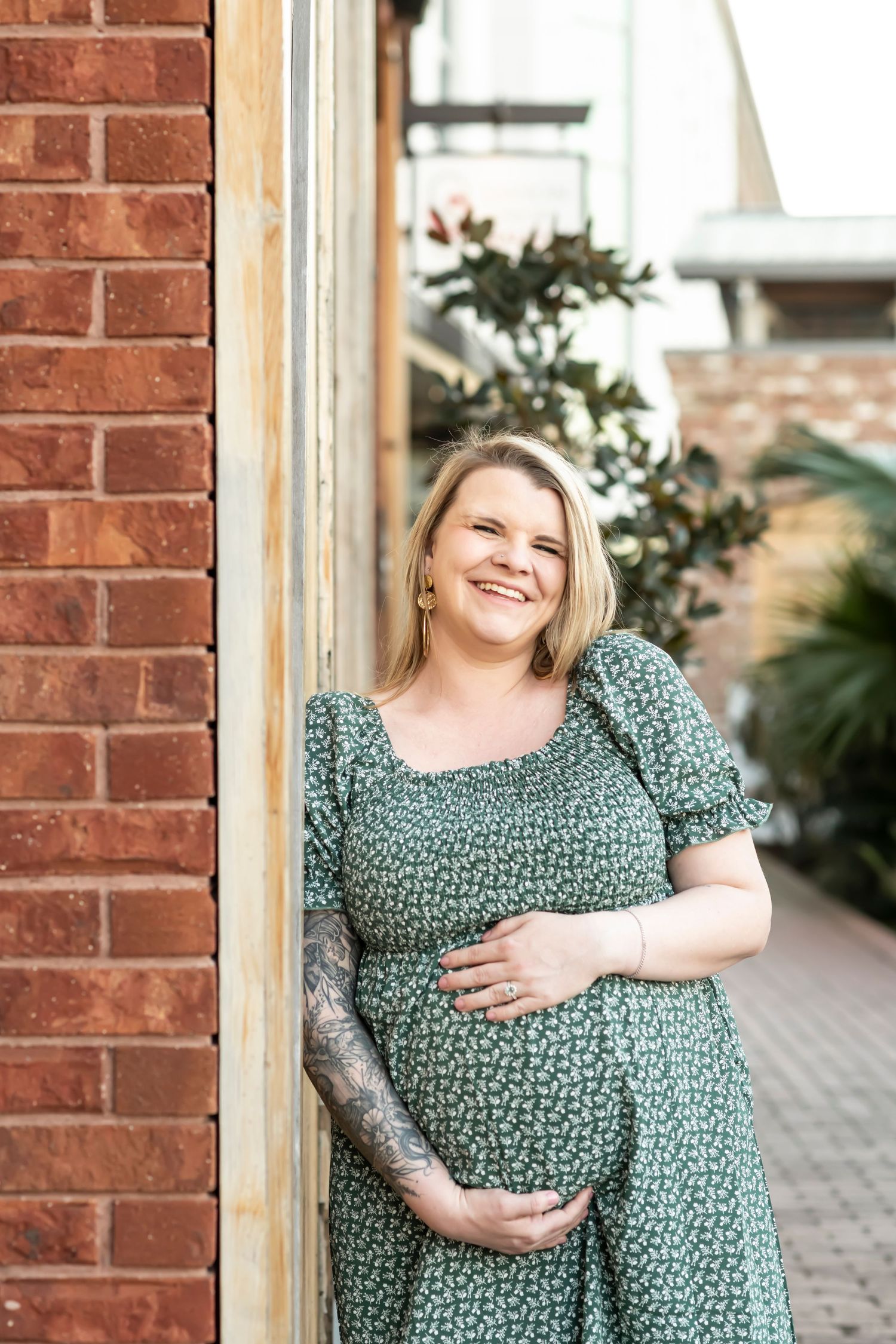 A pregnant woman in a green floral dress stands smiling beside a brick wall with plants in the background.