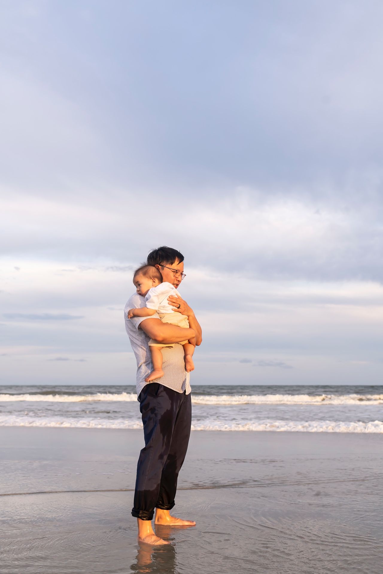 A person stands in casual attire holding another on an empty beach at sunset as waves roll in on the shoreline.
