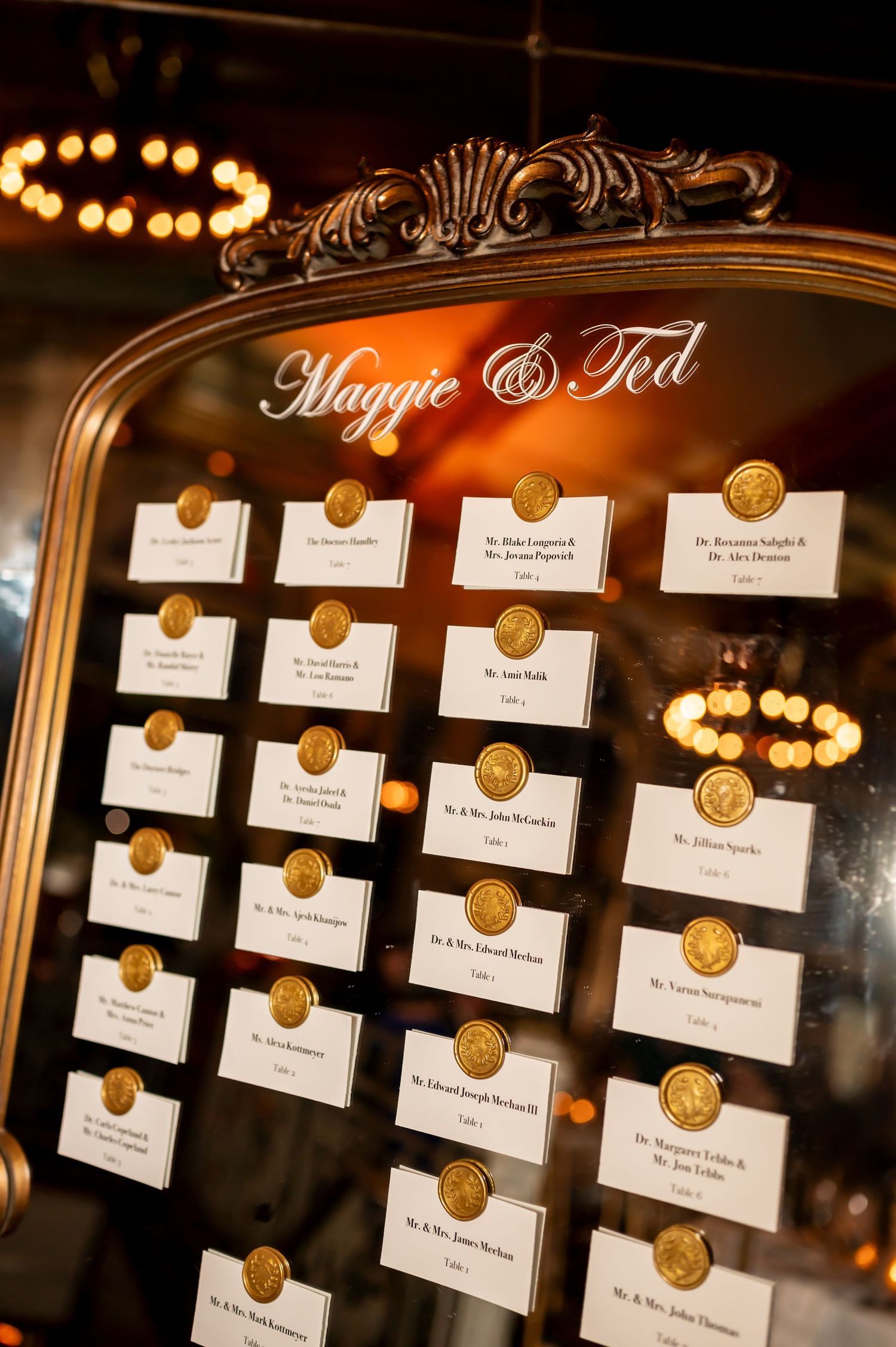 Elegant seating chart display with gold wax seals and names on white cards in an ornate vintage mirror frame reads 'Maggie & Ted'.