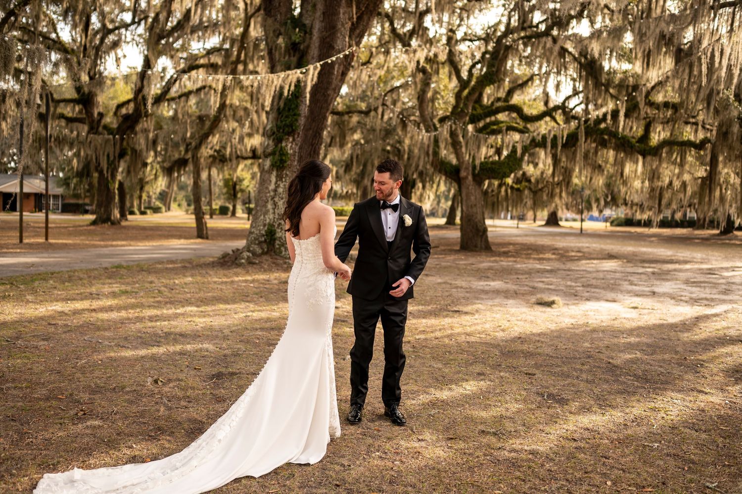 A couple in wedding attire stands together on a path under moss-draped oak trees during golden hour.
