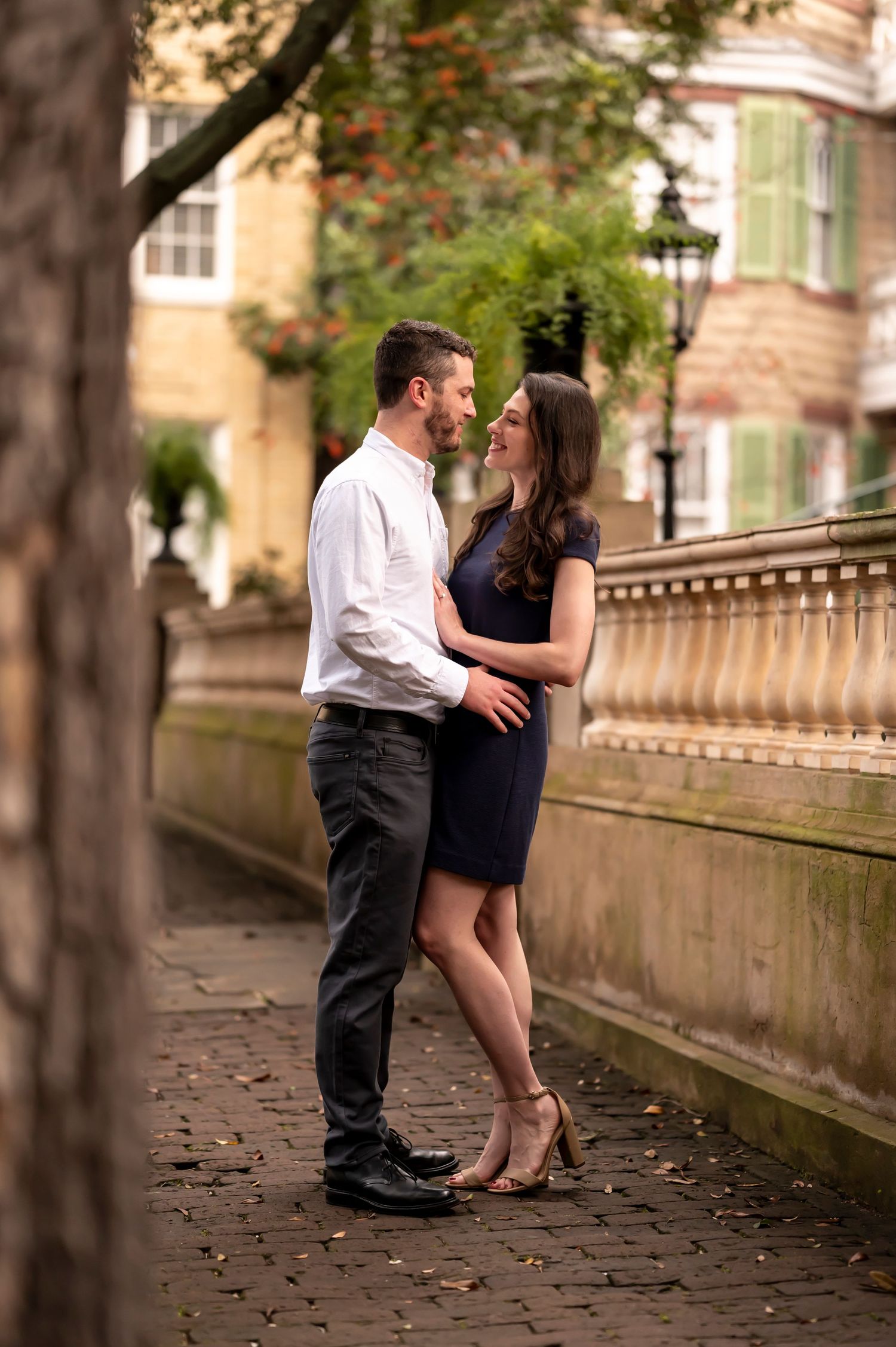 A couple shares a tender moment together on a stone pathway lined with trees and an ornate railing at dusk.