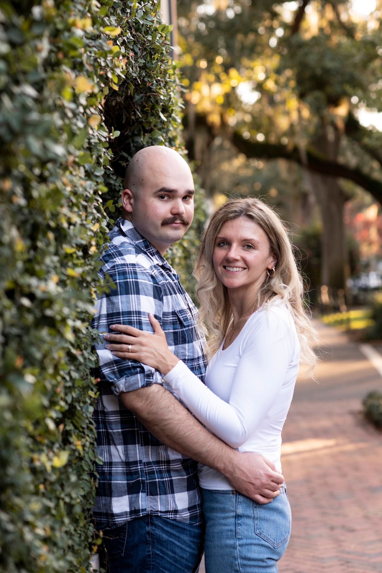A couple embraces against a leafy garden wall at sunset on a brick pathway during an engagement photo session.