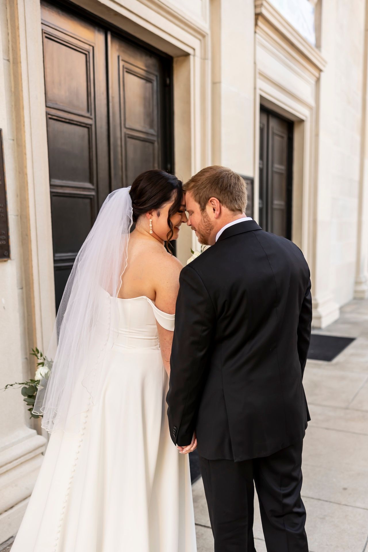 Newlywed couple shares intimate moment outside church doors in white wedding dress and black suit.