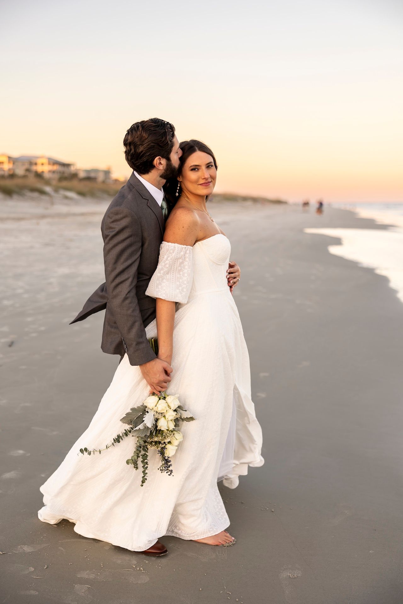 A romantic beach wedding photoshoot at sunset with couple in elegant white dress and dark suit on sandy shore.