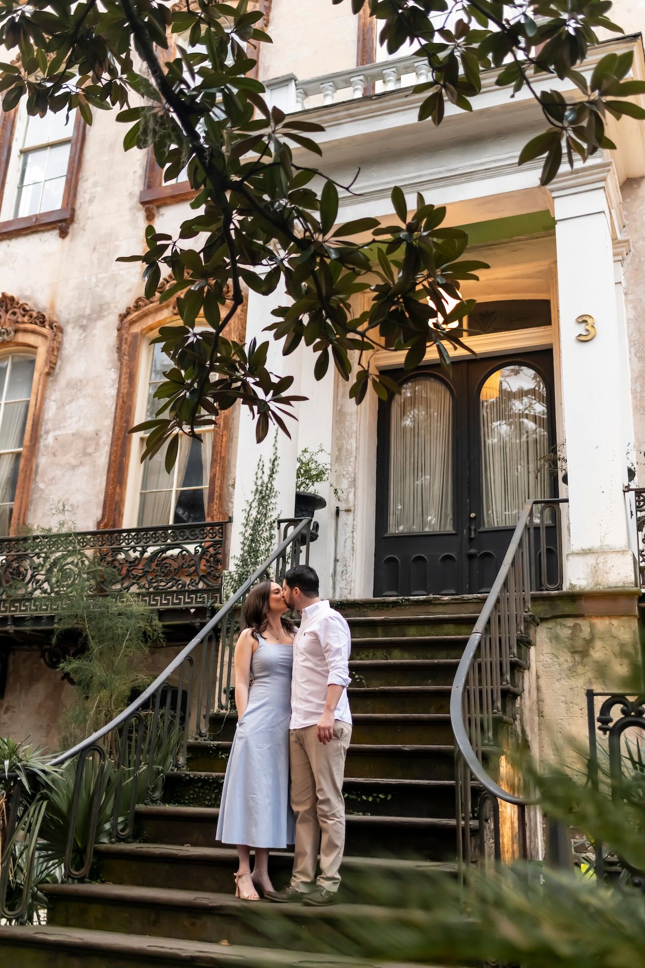 A couple stands on historic brownstone steps framed by magnolia leaves and iron railings in a romantic urban setting.