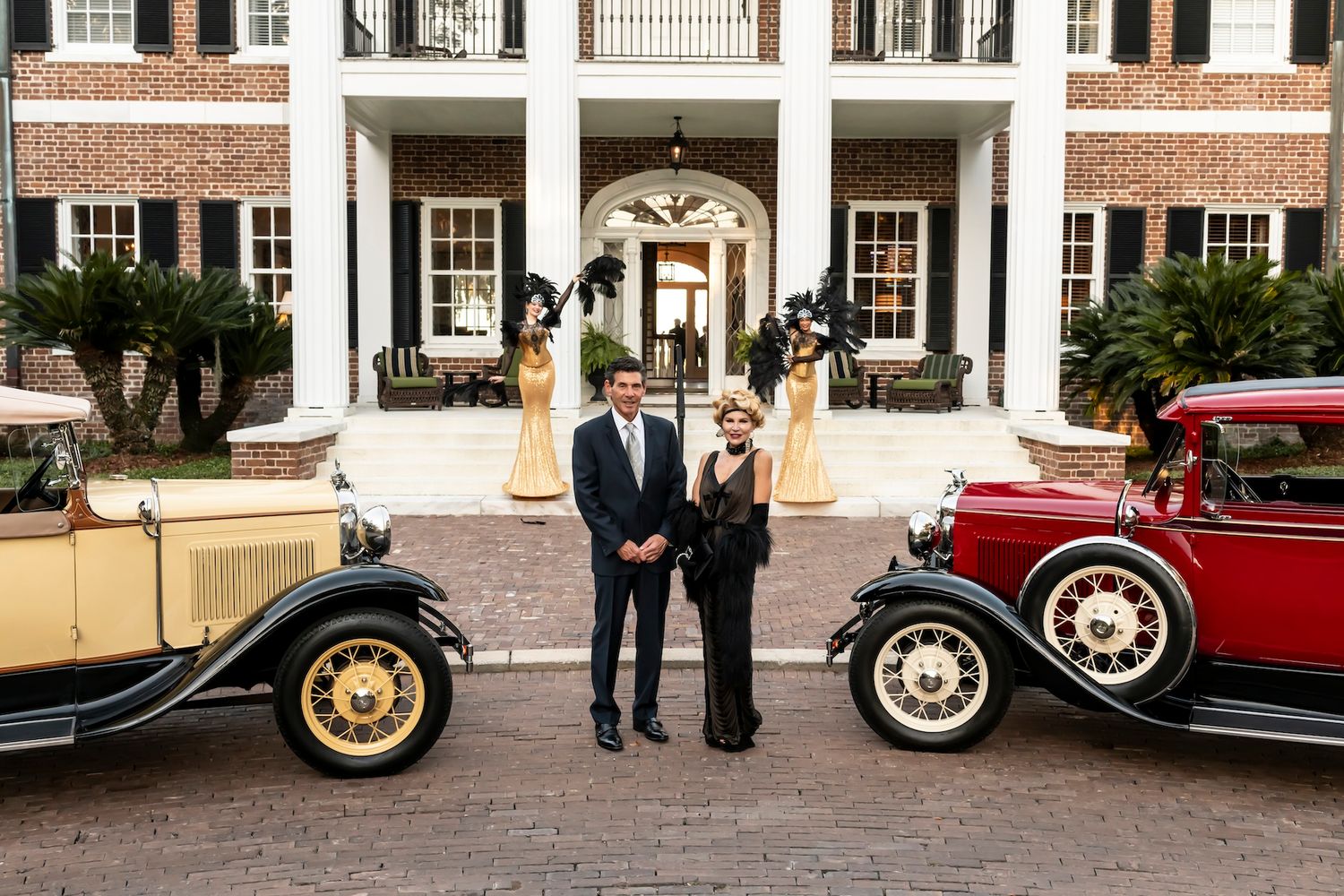 People in formal attire pose between two vintage automobiles outside a grand colonial-style mansion with white columns.