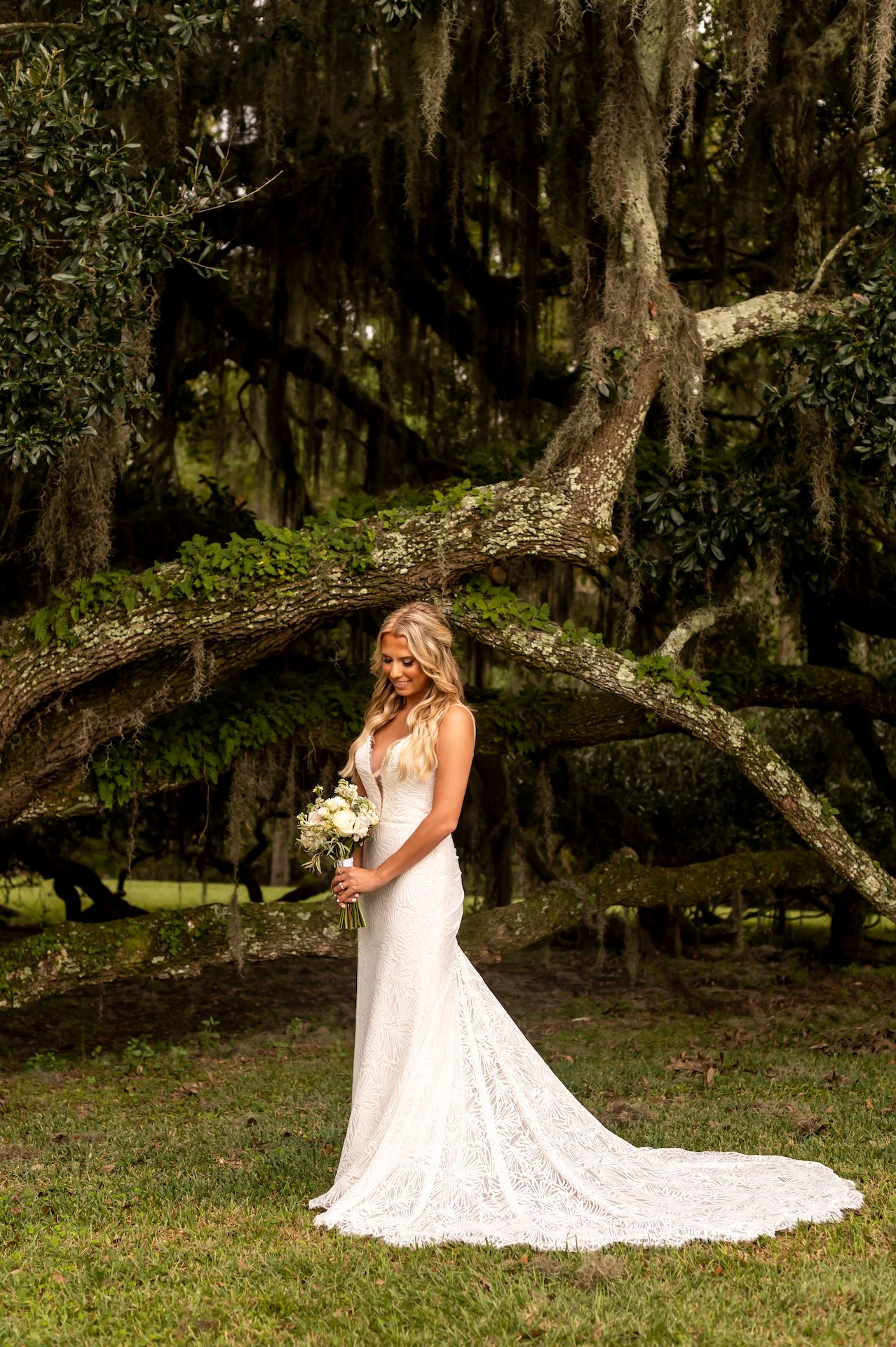 A bride in a flowing white lace gown poses elegantly in a Spanish moss-draped garden setting.