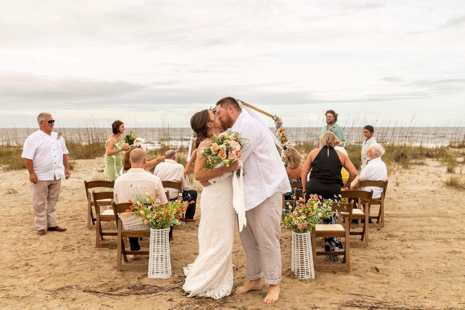 Newlyweds share intimate moment during beach ceremony surrounded by wooden chairs and floral arrangements.