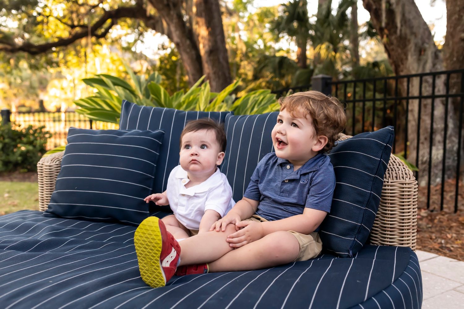 Two young children sit on a navy striped outdoor couch with throw pillows while enjoying a sunny afternoon.