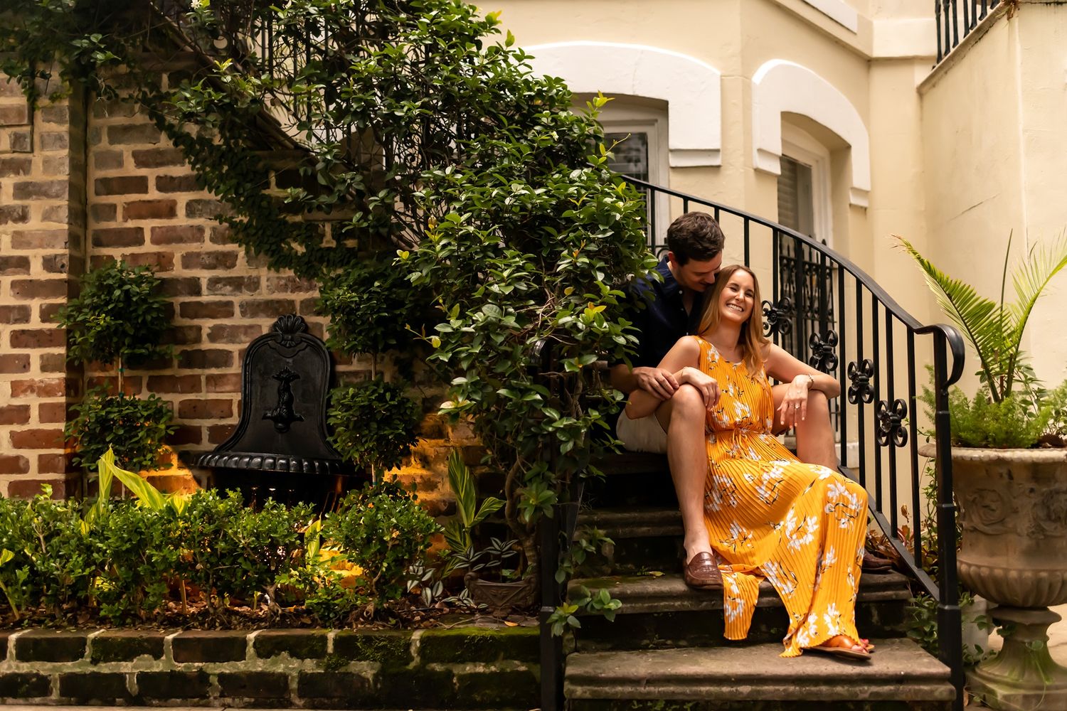 A person in a bright yellow dress sits outdoors on stone steps next to brick walls and lush greenery.
