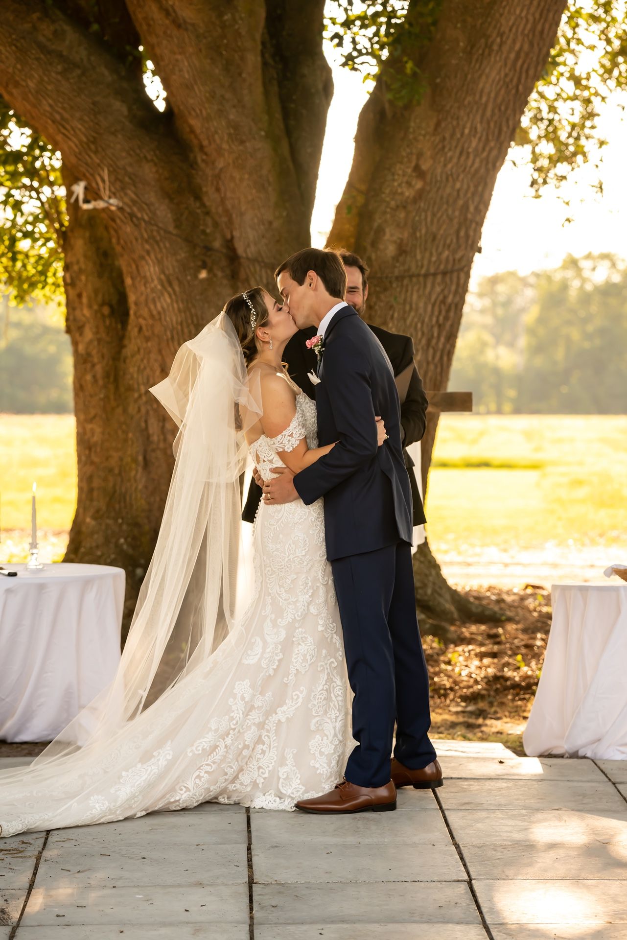 A couple shares an intimate kiss under sunlit oak trees during their outdoor wedding ceremony.