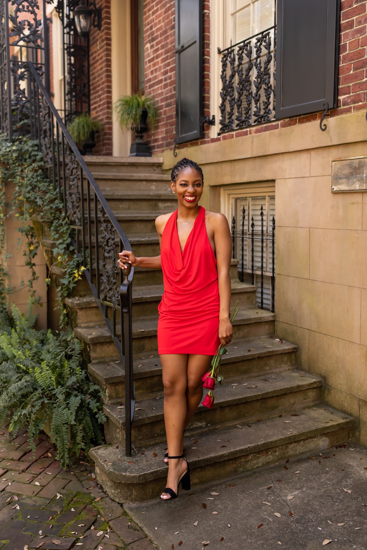 A woman in a vibrant red cocktail dress and heels poses on brownstone steps with ornate iron railings.
