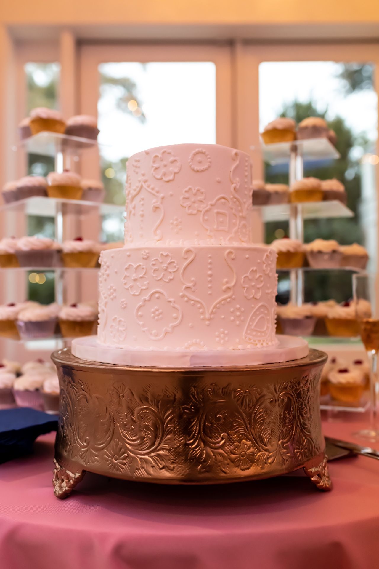 An elegant white wedding cake with delicate floral embossing sits on an ornate silver stand atop a pink tablecloth.