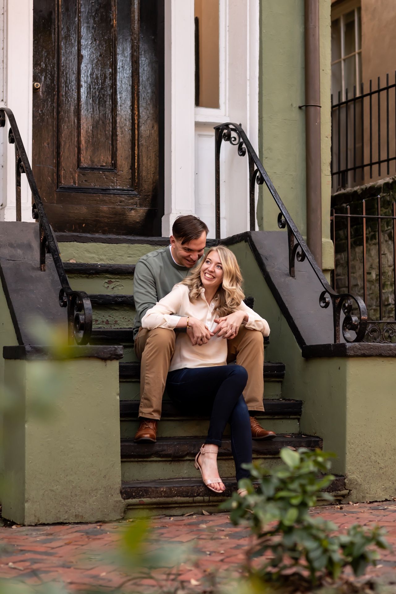 A couple sits close together on stone steps outside a historic building with ornate railings and a dark wooden door.