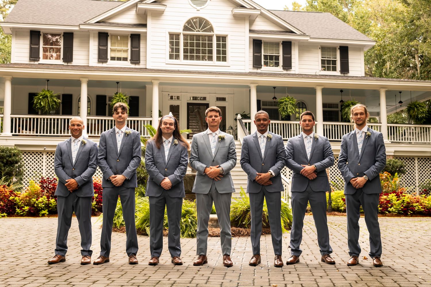 A group in matching gray suits stands in a line on a brick courtyard in front of a white colonial style mansion.