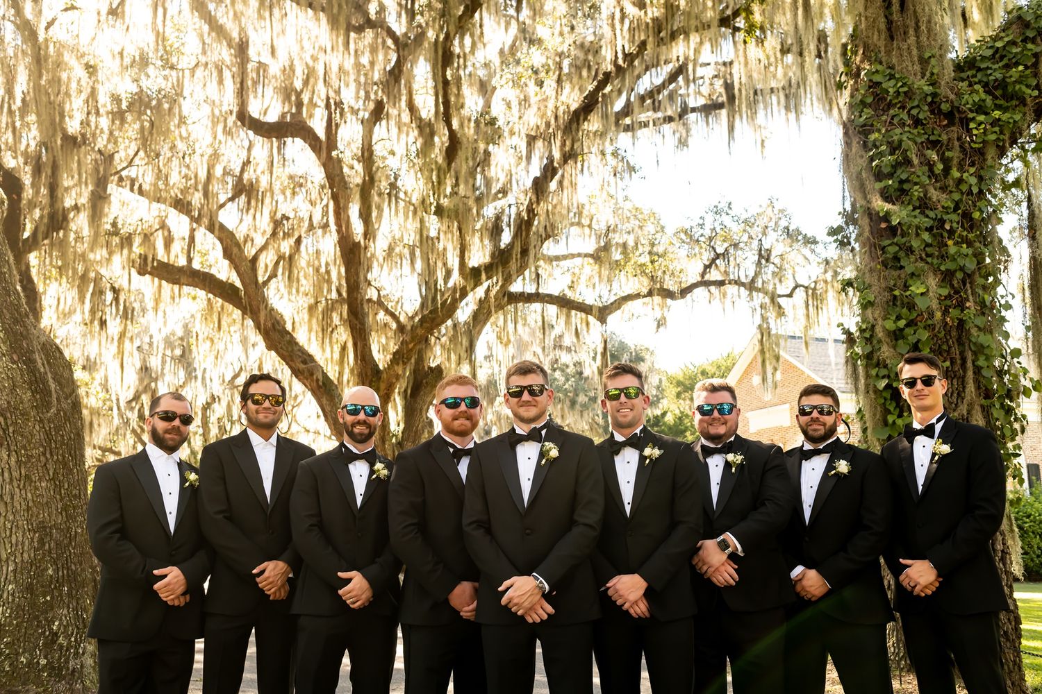 A formal wedding party in black tuxedos stands together under moss-draped oak trees in a Southern setting.