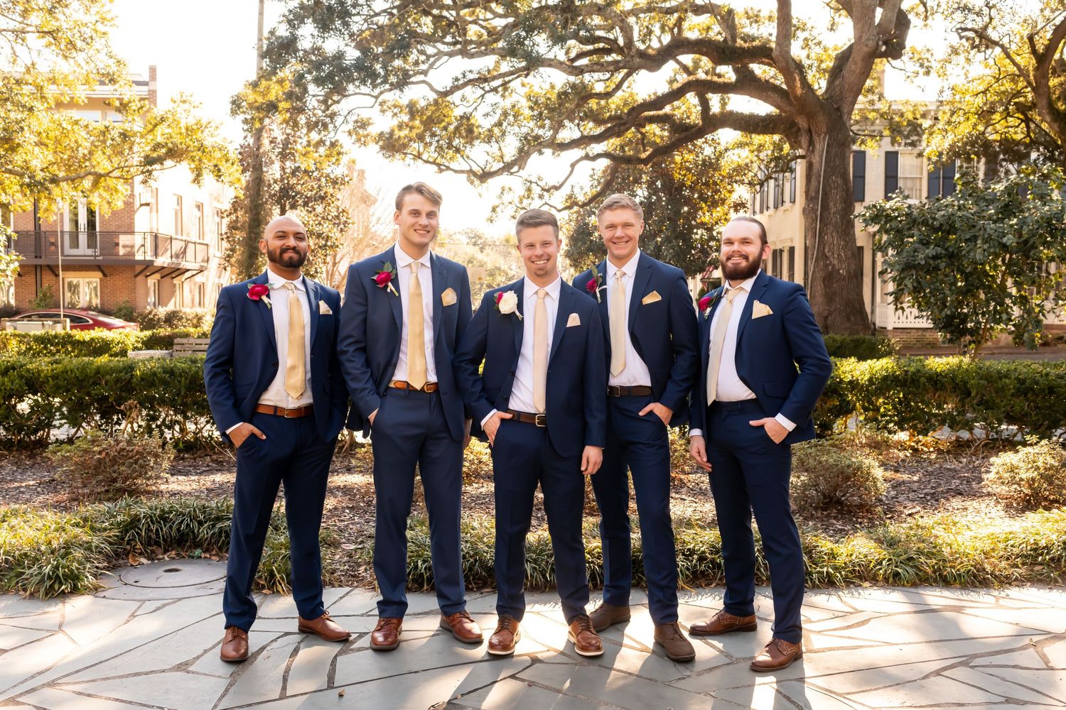 Wedding party groomsmen lined up outdoors wearing matching navy suits and posing casually in natural lighting.
