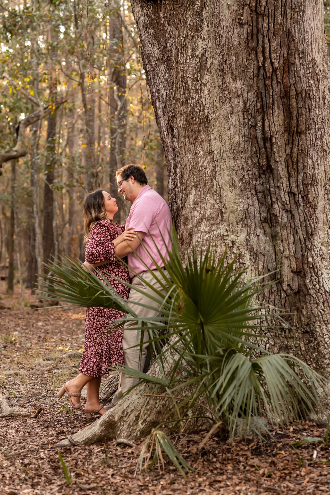 Romantic couple embraces near large oak tree in forest with palmetto plants during outdoor photoshoot.