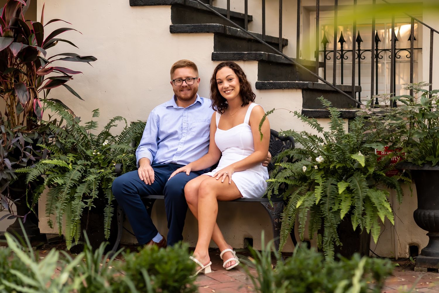A couple rests together on outdoor steps surrounded by lush ferns and greenery in a romantic setting.