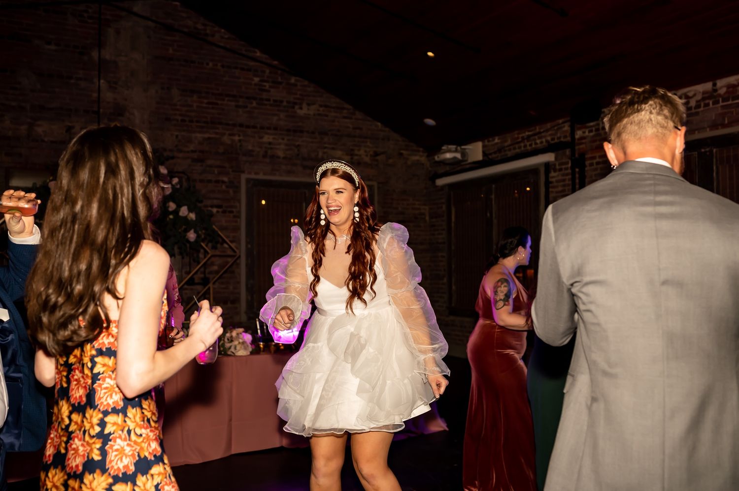 Wedding reception guests dancing and celebrating at a rustic-style indoor venue with warm lighting and brick walls.