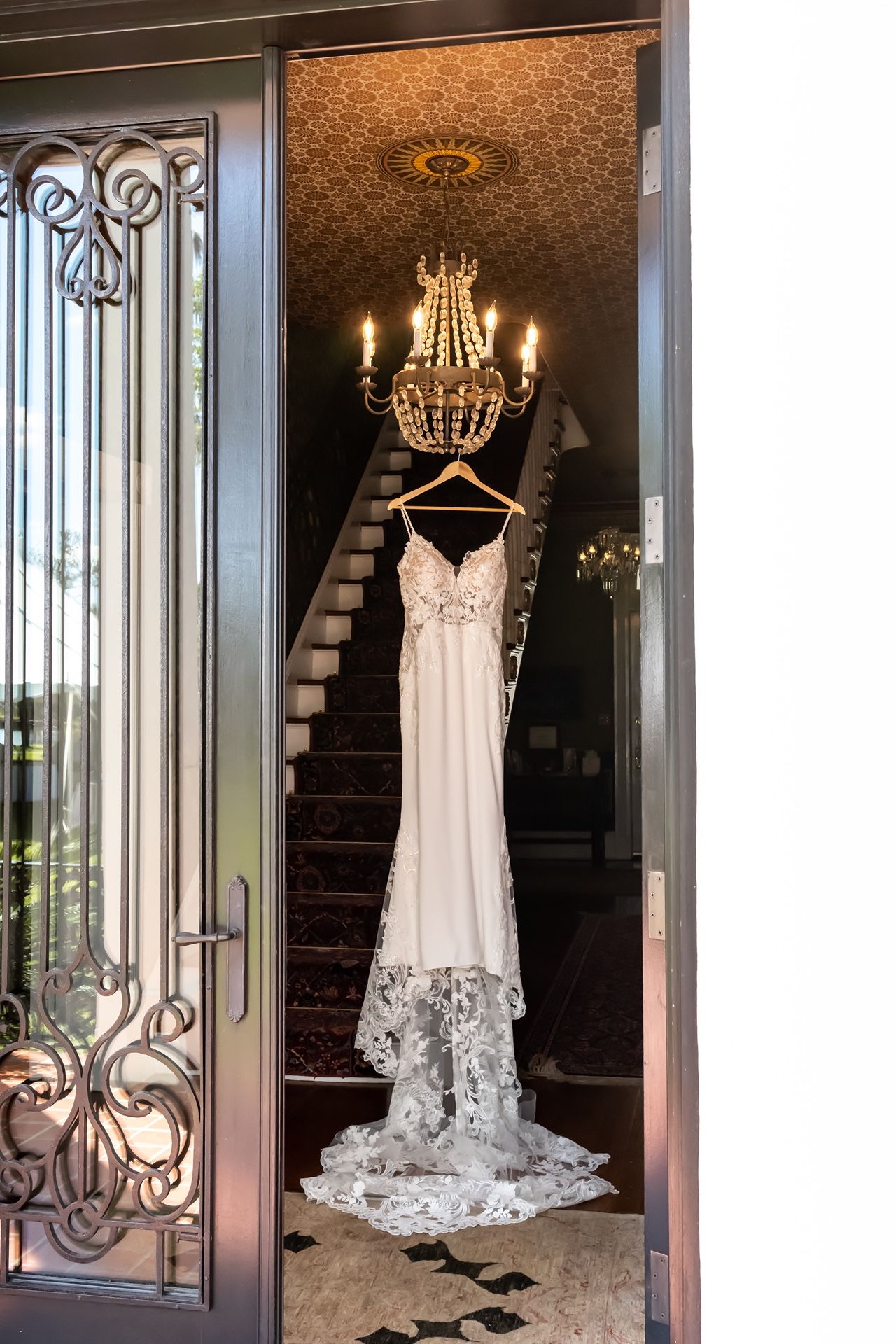 An elegant white lace wedding dress hangs in a doorway beneath a crystal chandelier with an ornate metal door and staircase visible.