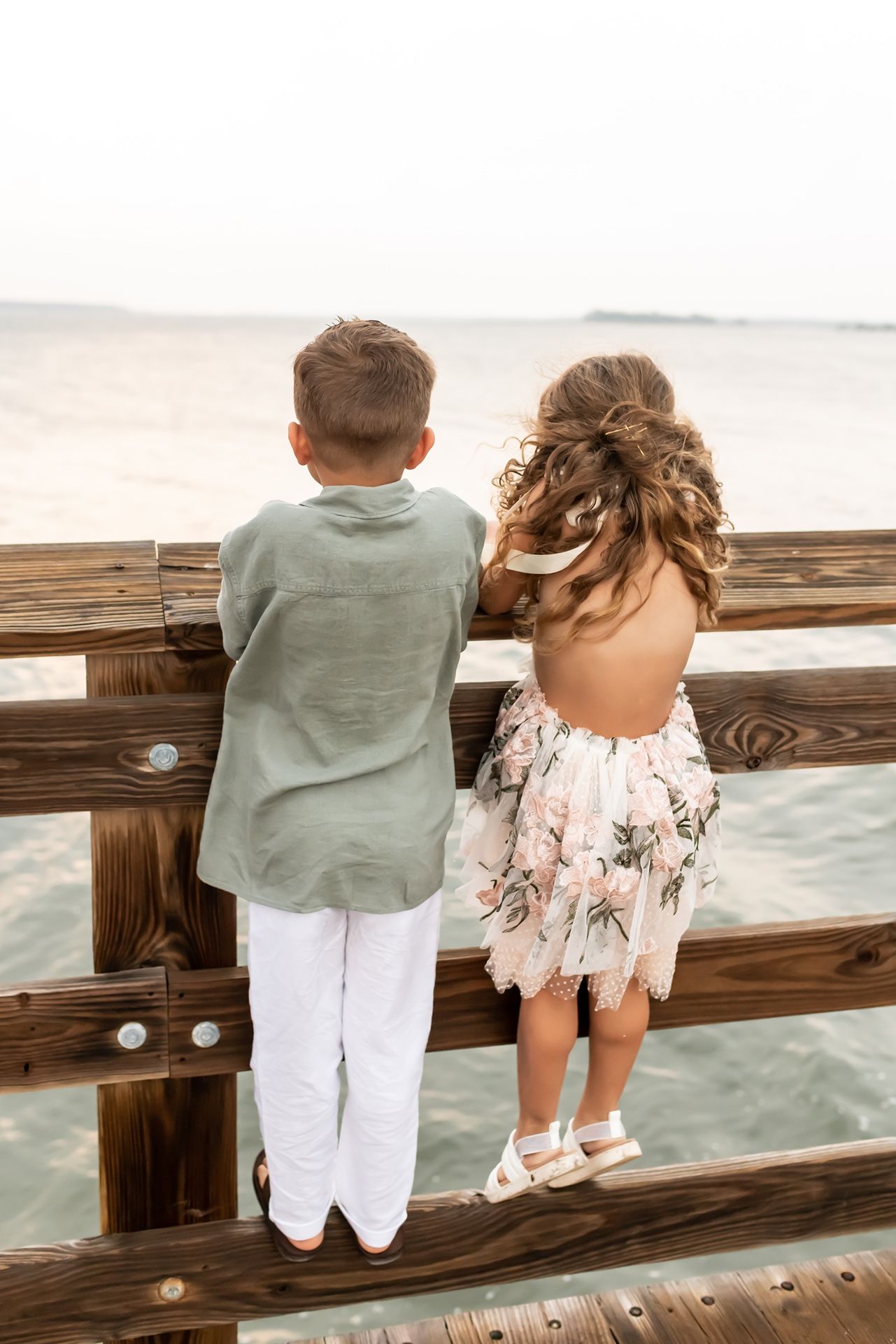 Two young children in summer outfits looking over a wooden pier railing at the ocean during sunset.