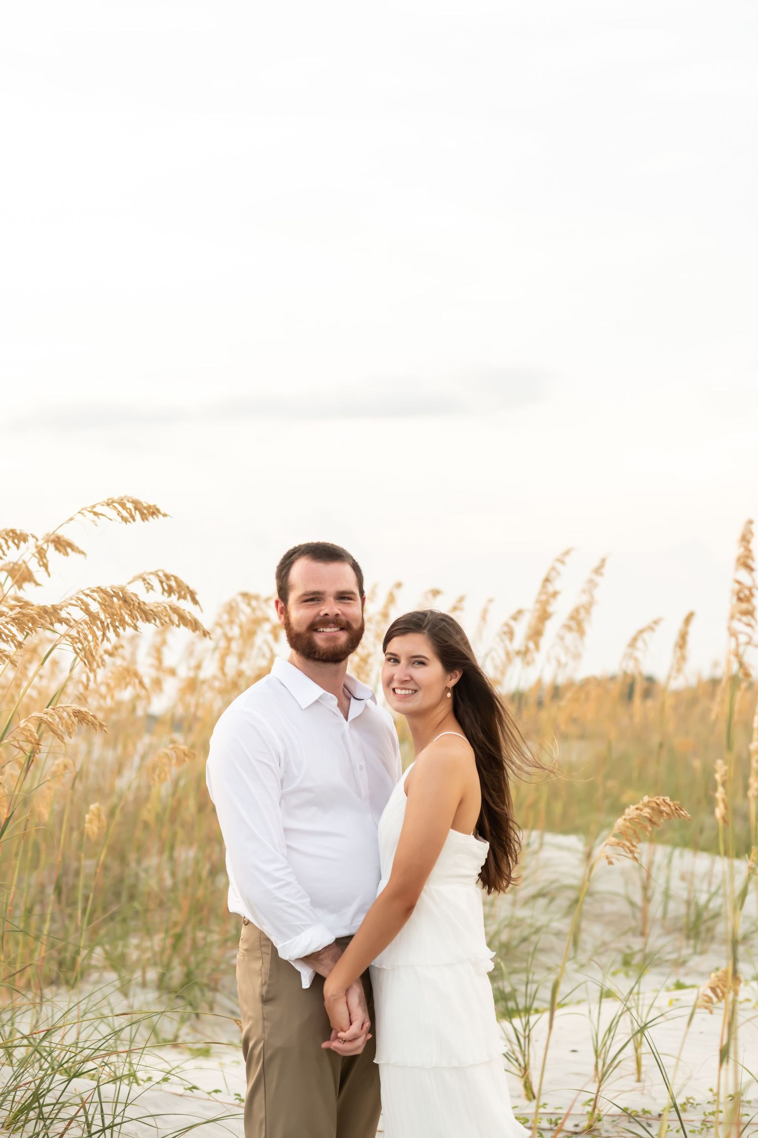 Couple embraces on sandy beach with sea oats swaying in golden sunset light.