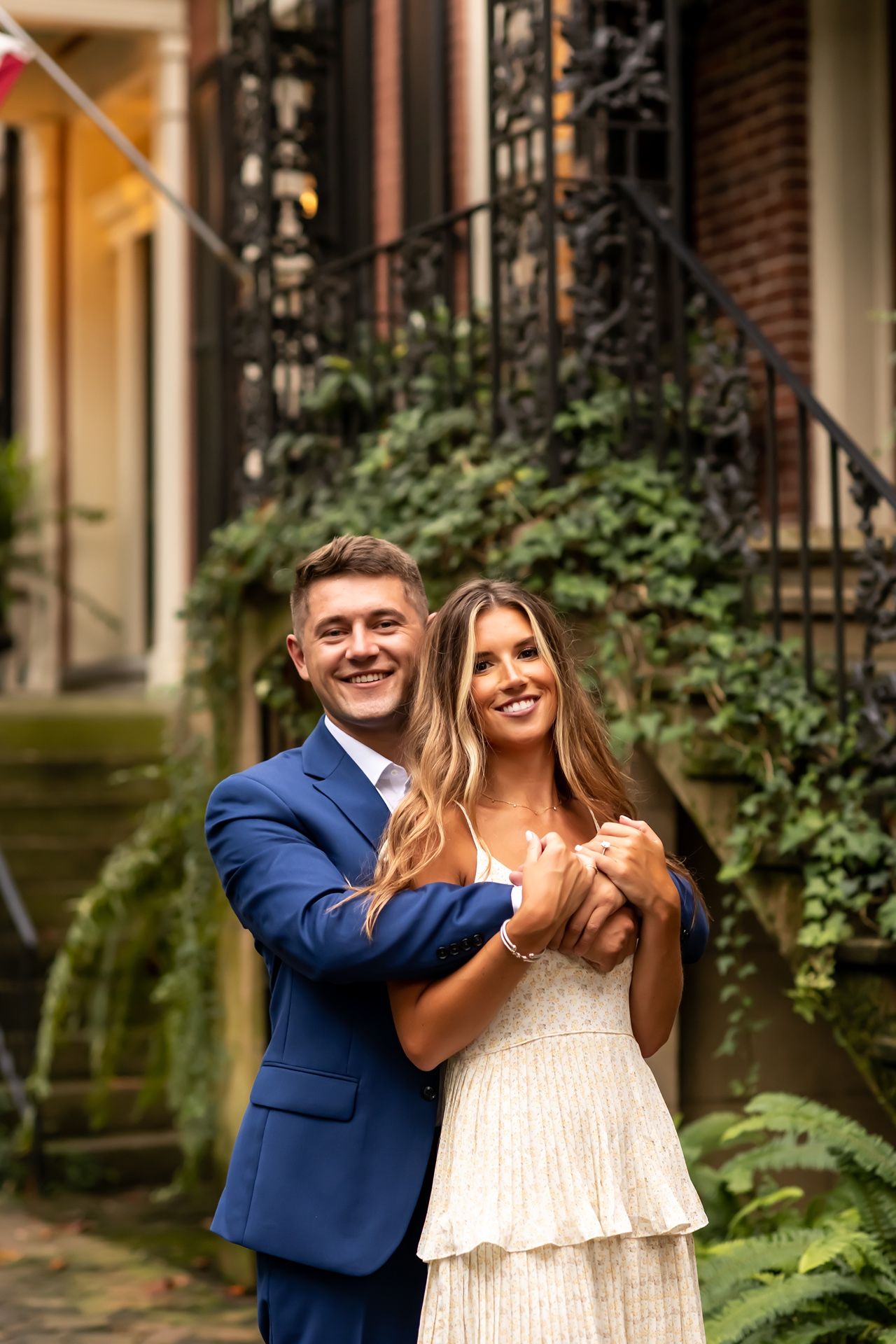 A couple celebrates their engagement with playful romantic poses on a brownstone porch with lush greenery.