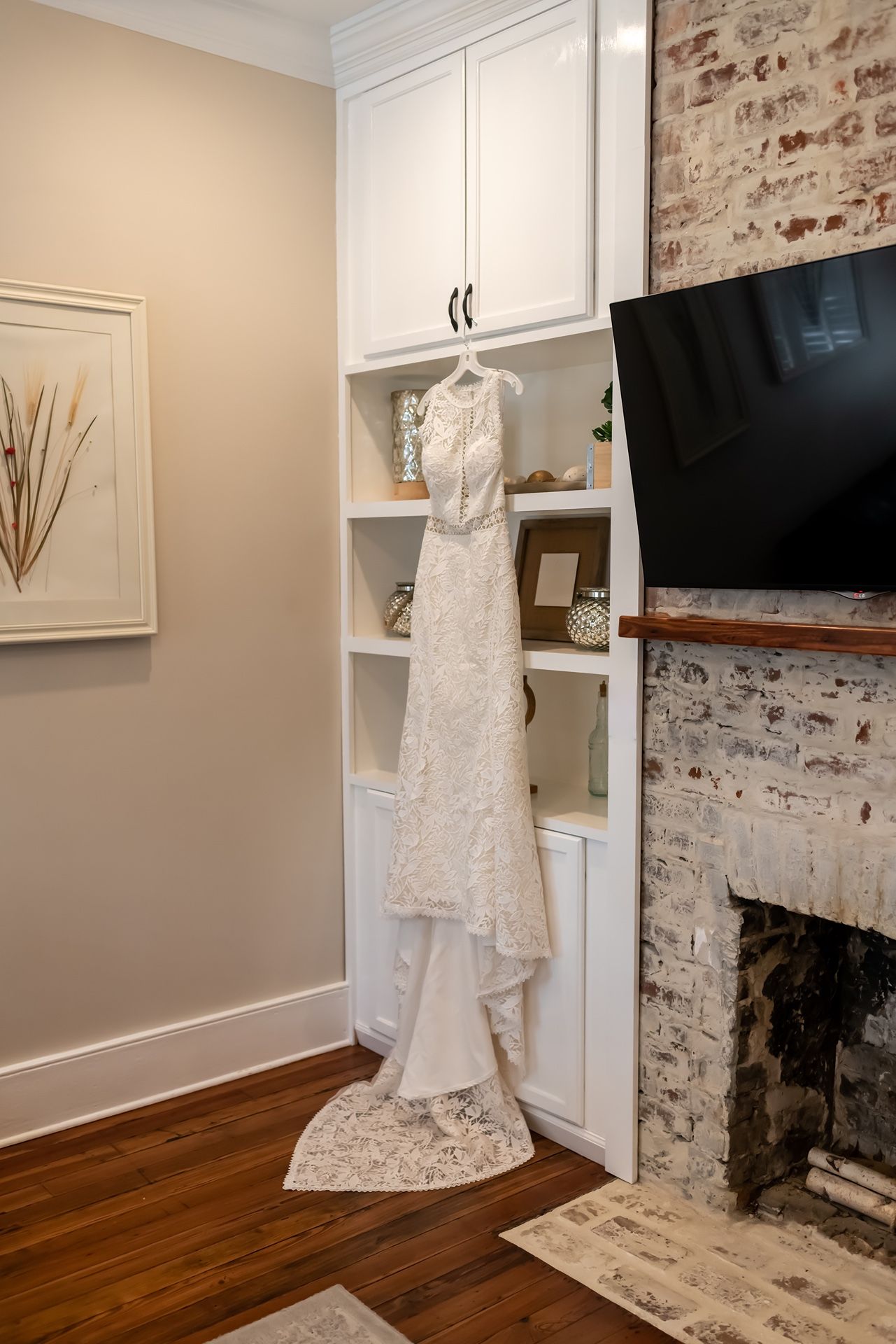 An elegant white lace wedding dress hangs on display next to a rustic brick fireplace in a cozy room with built-in shelving.