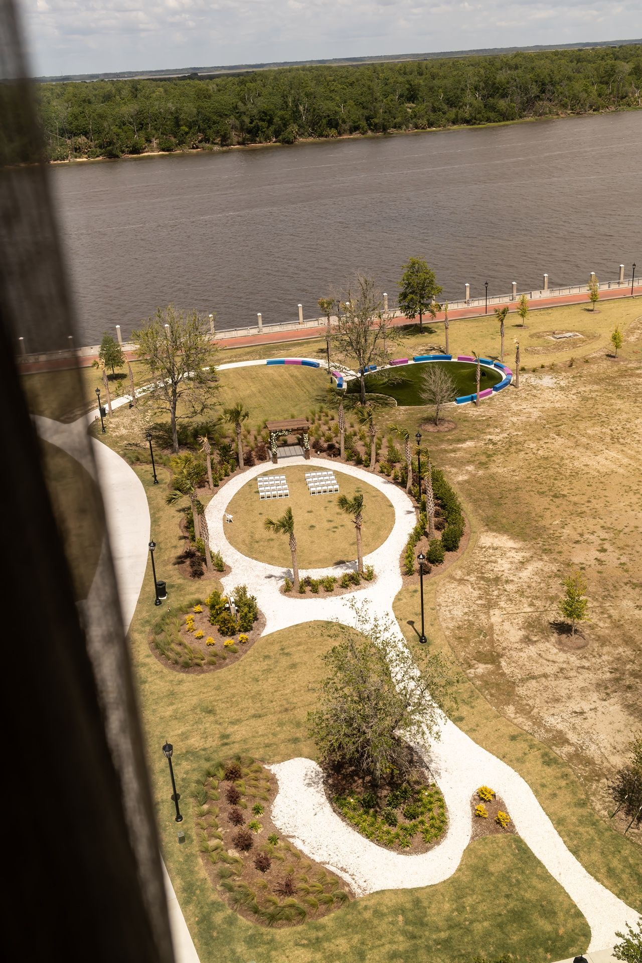 Aerial view of a curved walking path leading to a circular gathering area beside a river with landscaped gardens and trees.