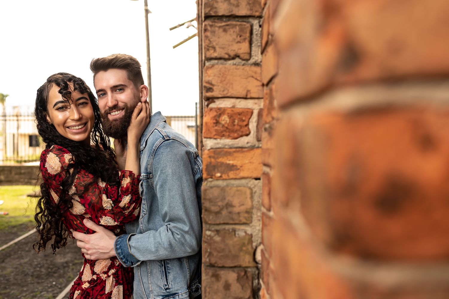 A couple embraces and smiles while standing next to a rustic brick wall in warm sunset lighting.
