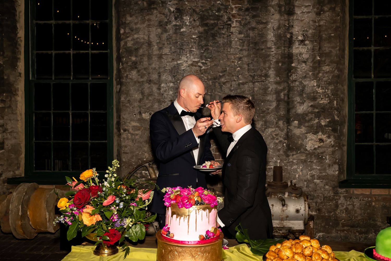 A couple sharing a romantic toast with wine glasses in front of a colorful cake and floral display at their rustic venue.