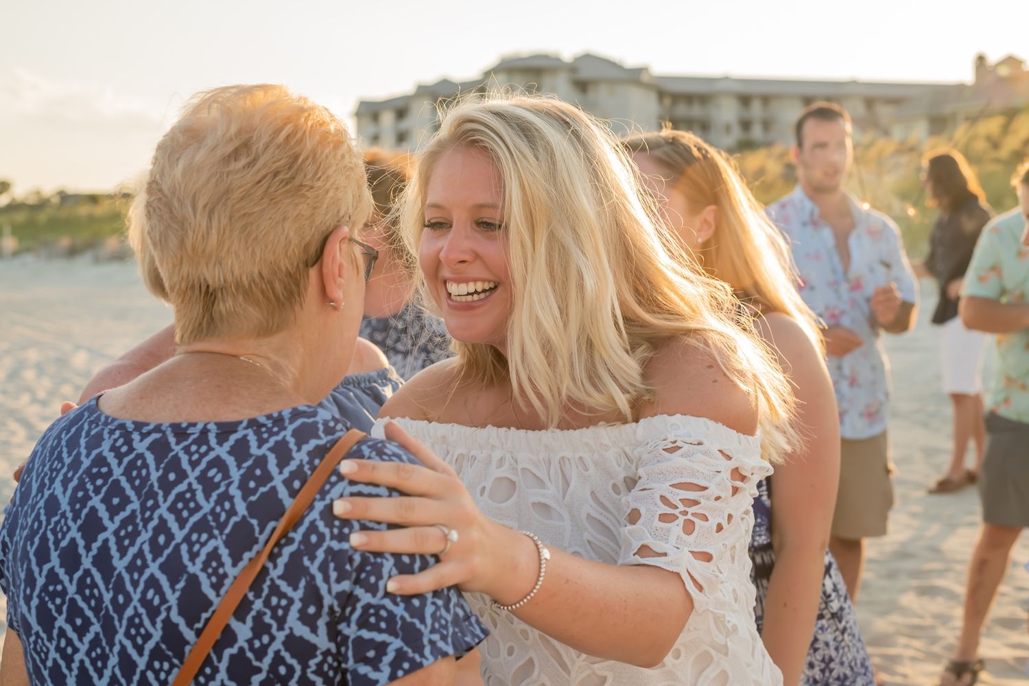 Two people embrace warmly on a sunny beach with others visible in the background during golden hour.