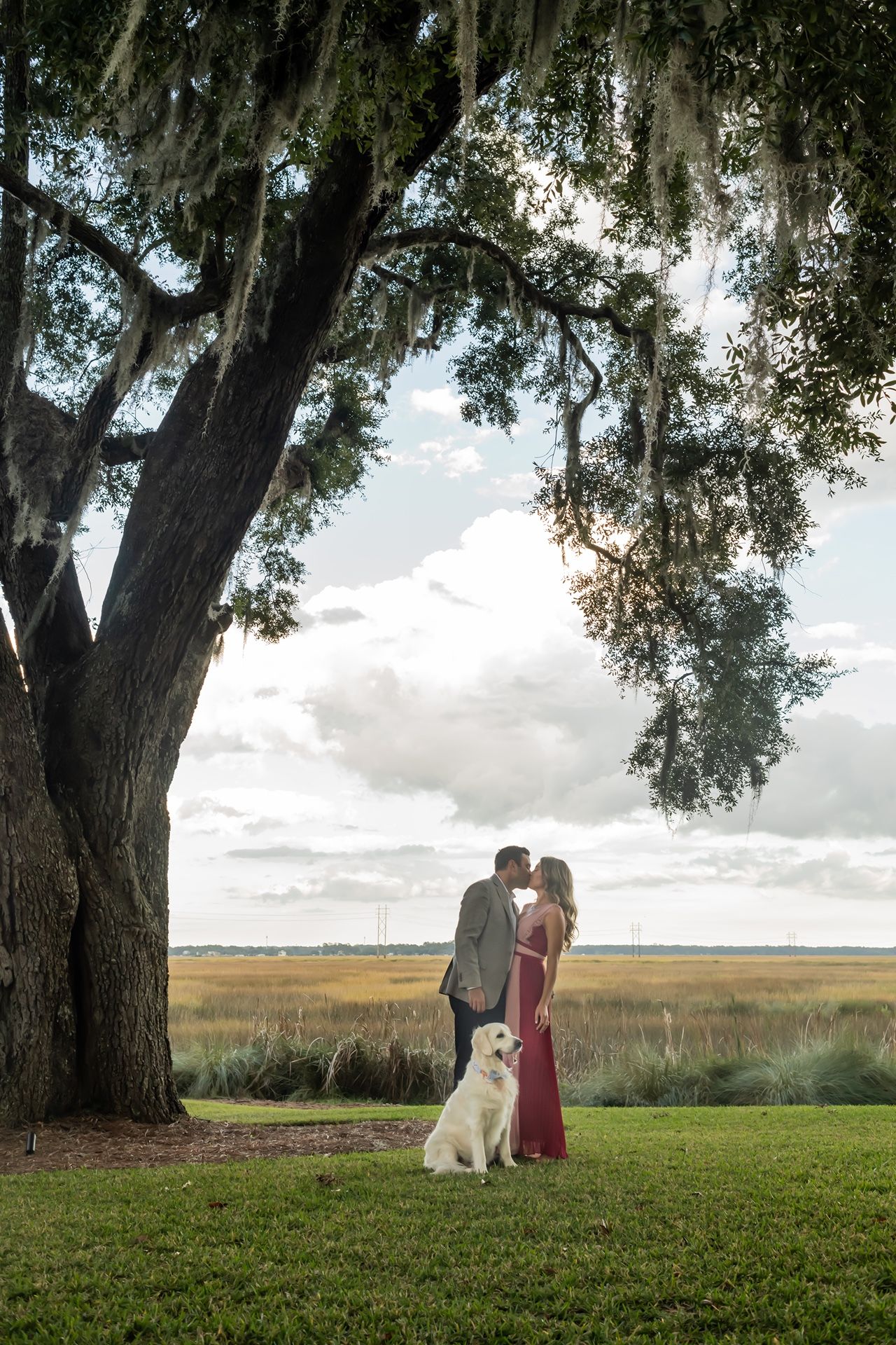 A couple stands together under a large oak tree with Spanish moss in a grassy marsh landscape at sunset.
