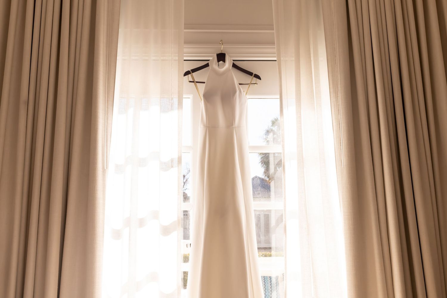 Wedding gown hanging in an elegant reception venue with cream-colored curtains and natural lighting.