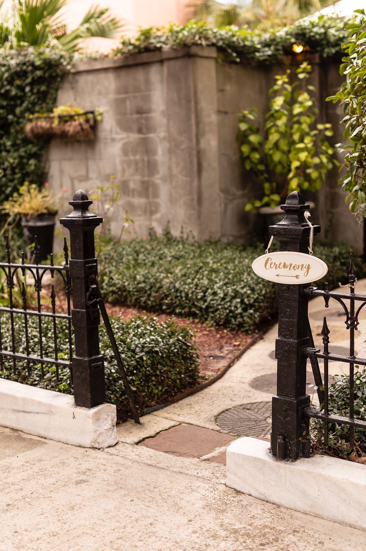 Black iron gate posts with decorative details frame a garden entrance with stone walls and lush greenery in Charleston.