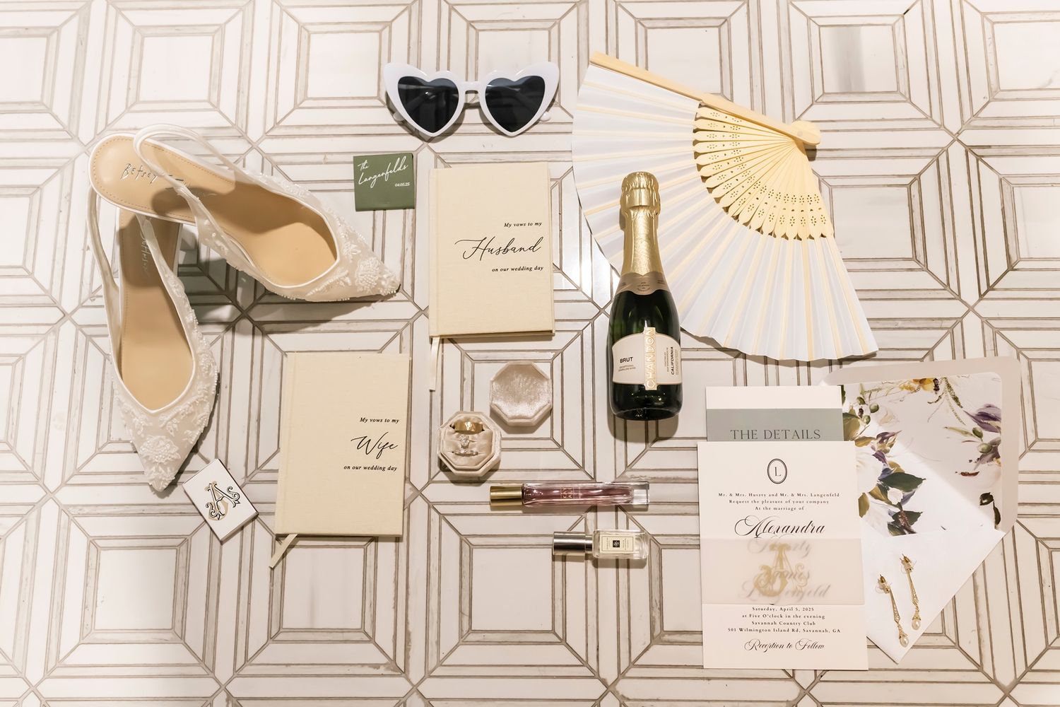 Flat lay of wedding accessories with champagne, heart sunglasses, shoes, and invitations on geometric patterned surface.