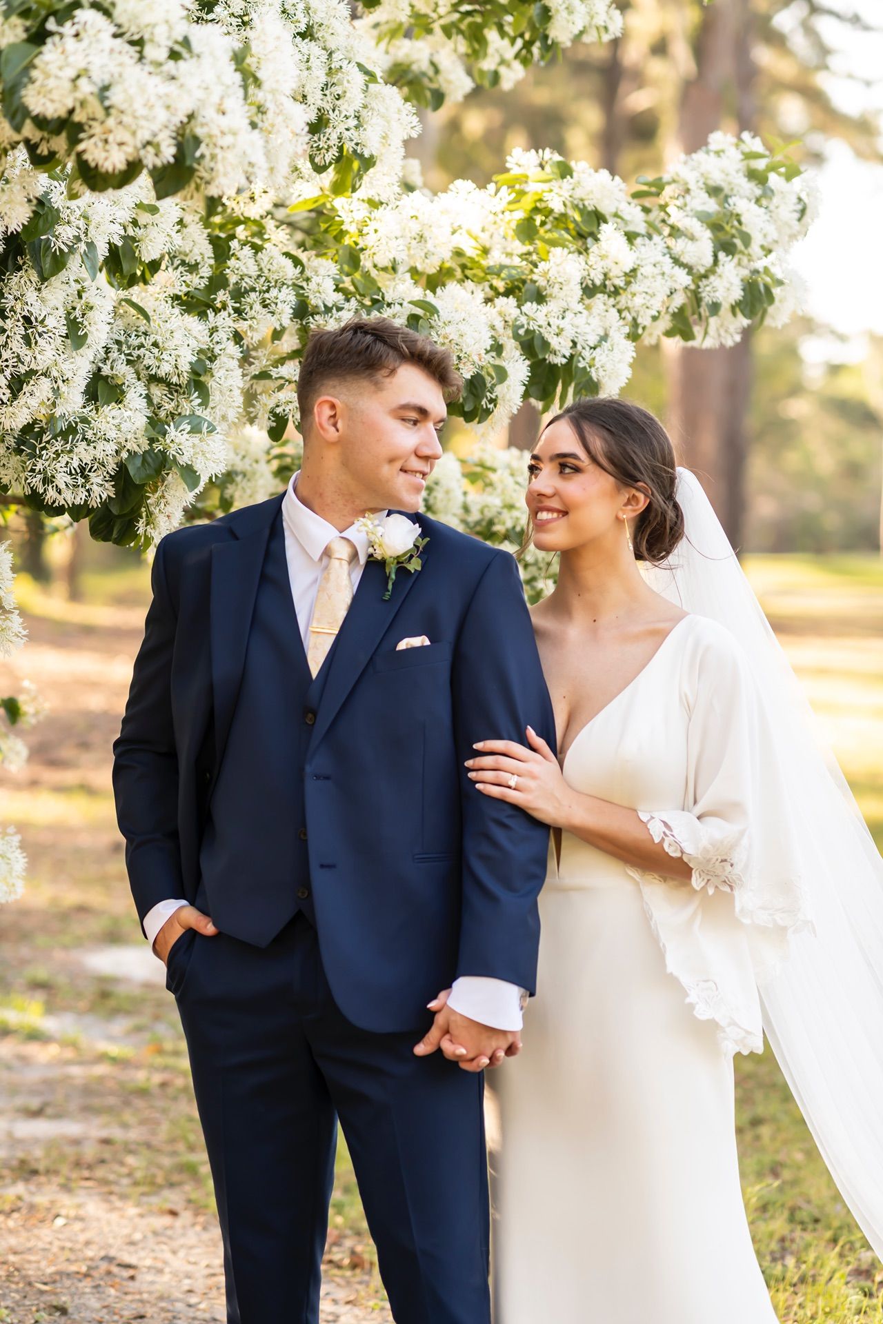 Elegant couple stands beneath white spring blossoms in romantic wedding portrait.