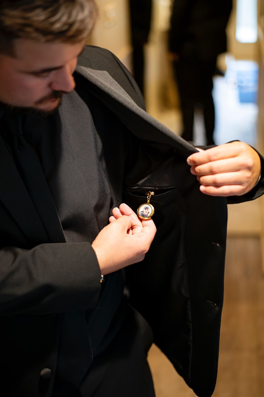 Someone adjusts a pocket watch while wearing a formal black suit jacket in an indoor setting.