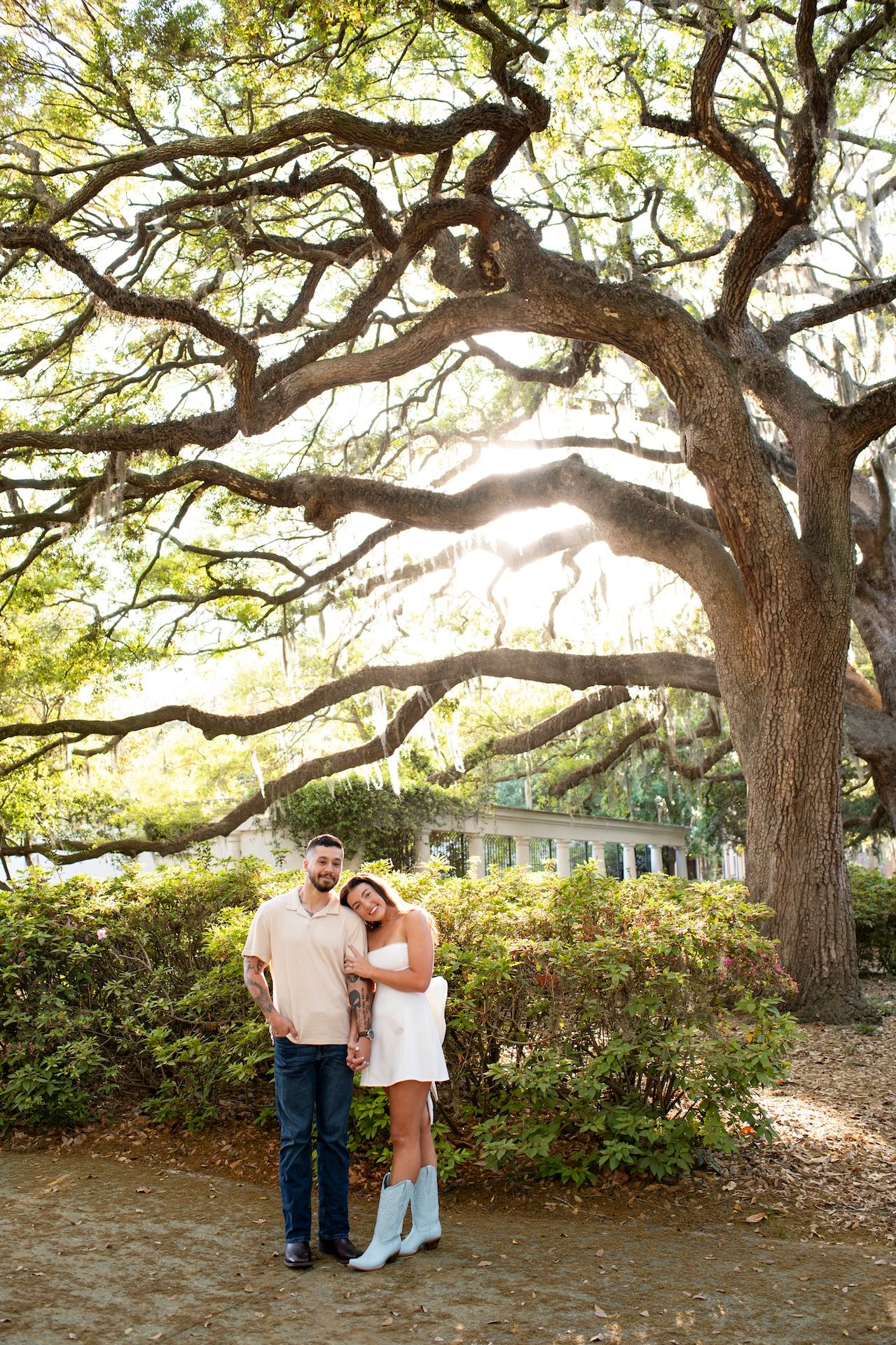 Couple posing under a large oak tree with sunlight filtering through branches in a lush garden setting.