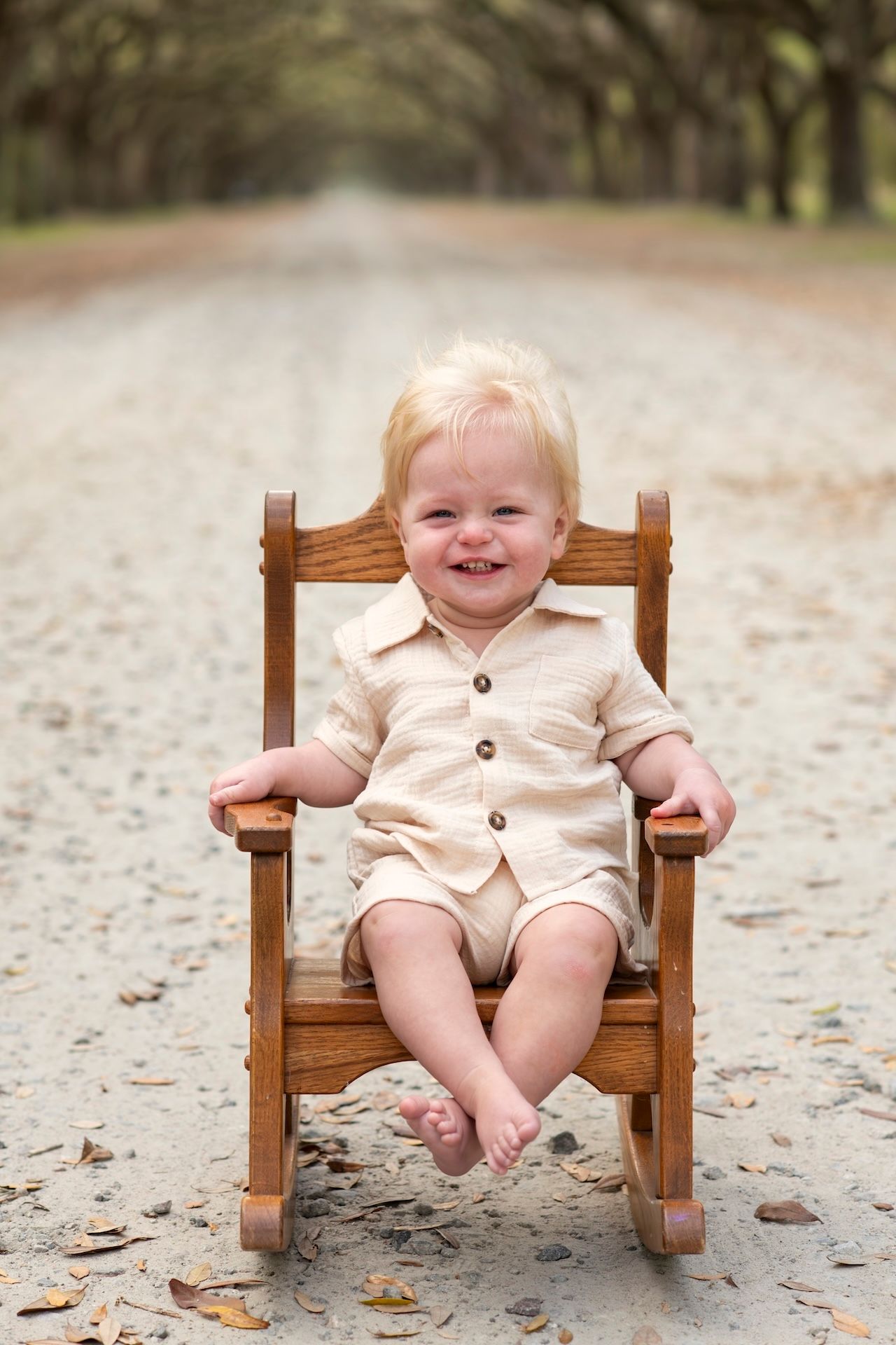 Smiling toddler in beige romper sitting in small wooden rocking chair on tree-lined dirt road.