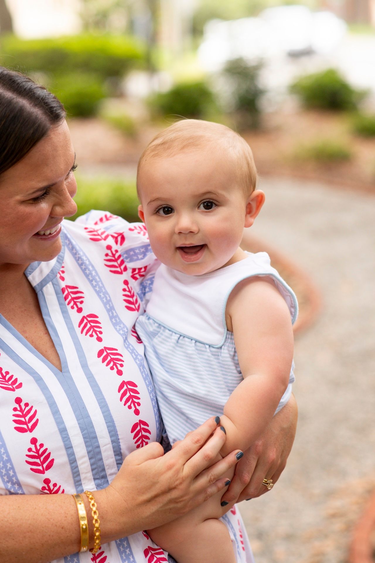 A smiling woman in a blue striped dress holds a happy baby girl in a light blue dress outdoors.