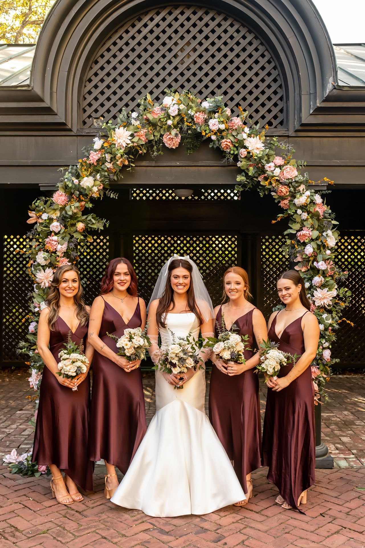 Bride in white gown poses with bridesmaids in burgundy dresses under a floral arch at outdoor venue.