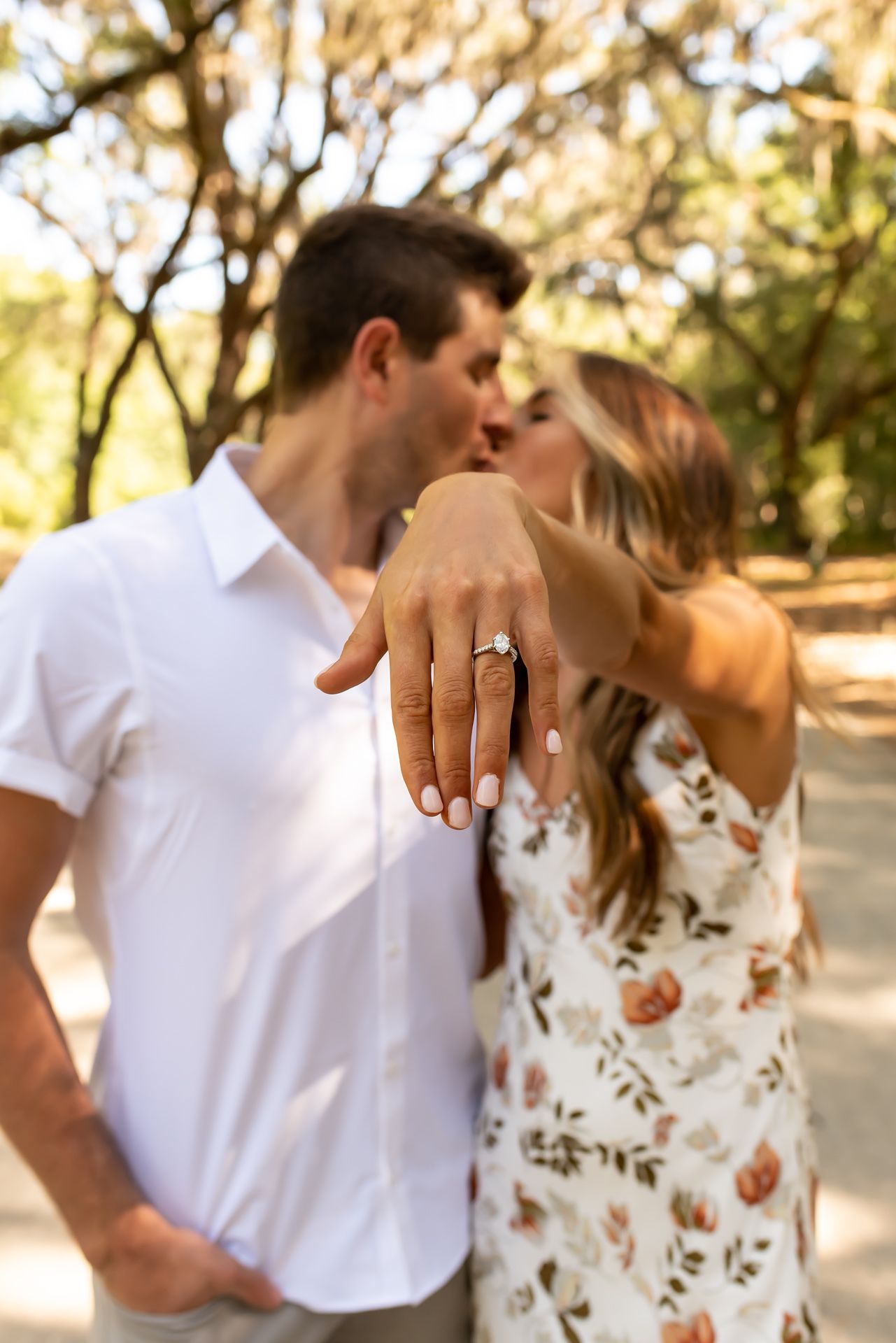 A couple wearing white and floral outfits embracing outdoors in a garden setting while showing off a ring.