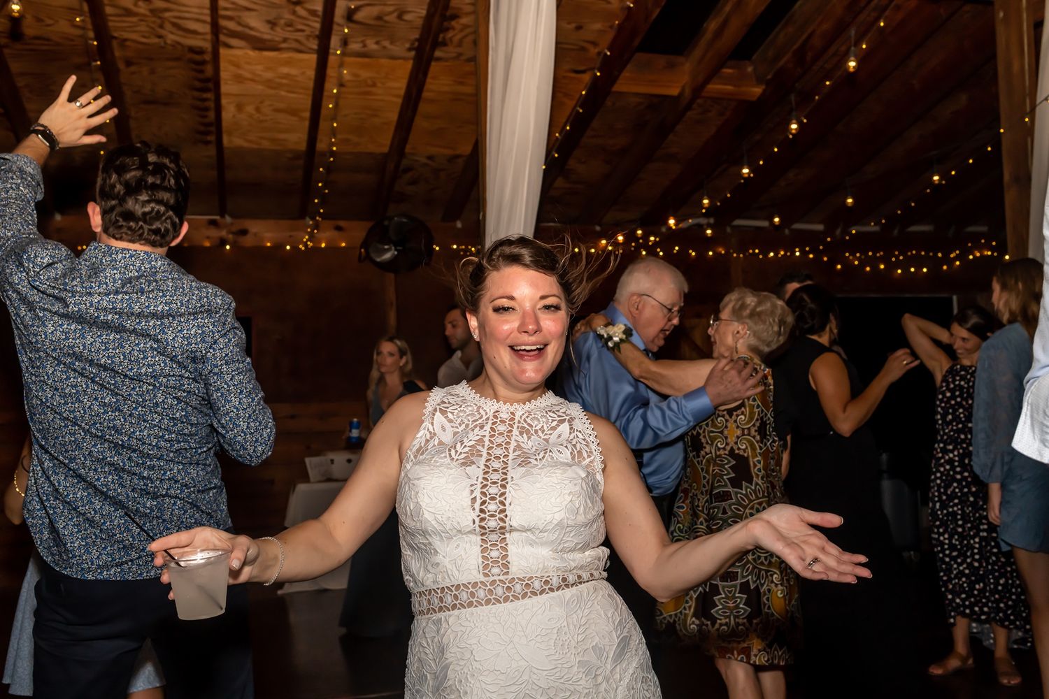 People dancing and celebrating at a rustic reception venue with string lights and wooden beams overhead.