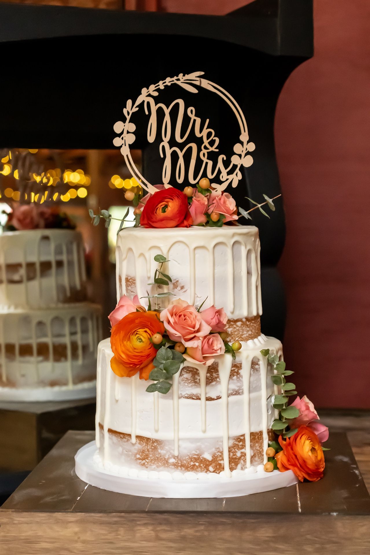 Elegant two-tier white wedding cake with dripping icing, coral roses, and a gold Mr & Mrs cake topper.
