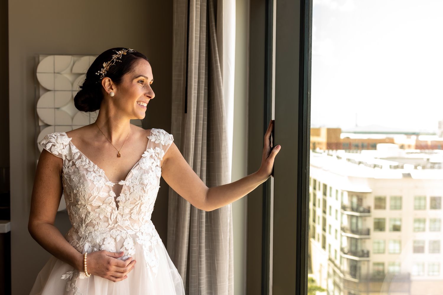 A person in an elegant white wedding gown gazes out a window while holding the curtain.