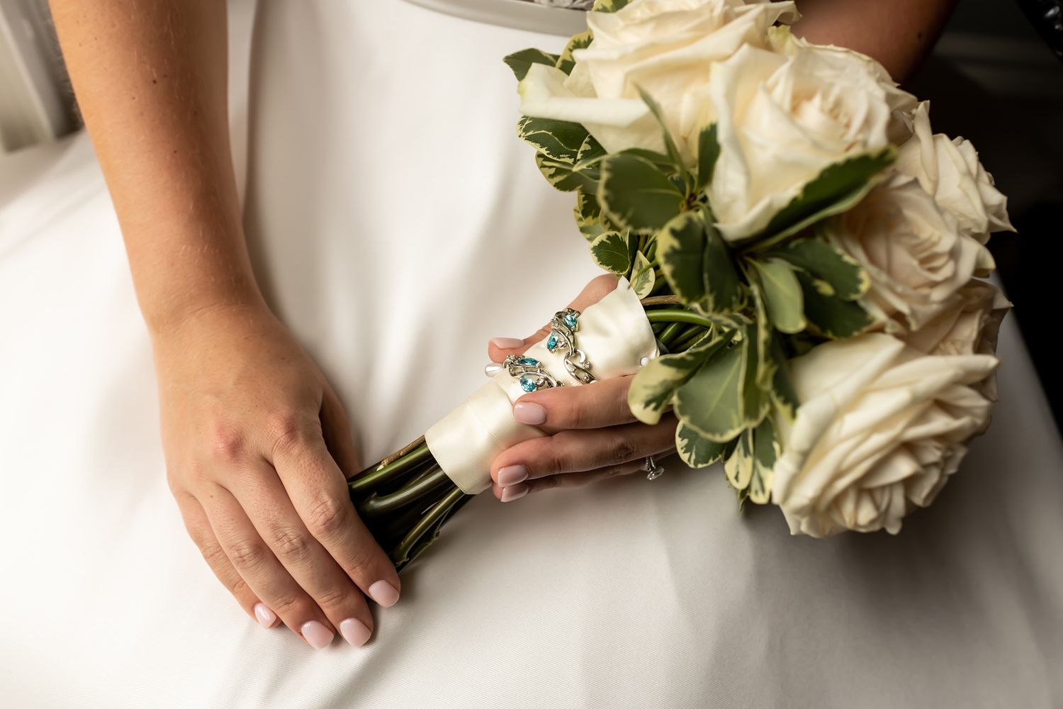 A white wedding bouquet with cream roses wrapped in lace trim rests on a white dress fabric.